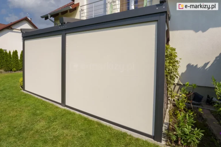 Dwa refleksole Ziiip 120 Selt zamocowane do konstrukcji pergoli, w kolorze konstrukcji antracytowym, dopasowanym do barw zadaszenia. Beżowa tkanina chroni przed słońcem i niepożądanymi spojrzeniami. Przed roletami widoczny trawnik a obok pergoli kwietnik z zielonymi roślinami.