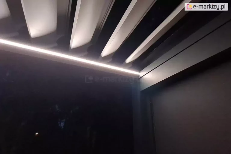 Zdjęcie prezentuje zbliżenie na detal wykończenia nowoczesnej pergoli tarasowej w warunkach nocnych, ze szczególnym uwzględnieniem systemu oświetlenia LED. W górnej części kadru widoczne są fragmenty aluminiowych lameli dachowych w jasnym kolorze, które są ustawione pod kątem, w pozycji półotwartej. Poniżej lameli, wzdłuż wewnętrznej krawędzi ciemnej, antracytowej ramy konstrukcyjnej, zamontowana jest liniowa listwa LED. Emituje ona intensywne, chłodne białe światło, które tworzy wyraźną, świetlistą linię oddzielającą dach od bocznej części pergoli. Po prawej stronie widoczny jest fragment masywnej ramy oraz górna część kasety rolety bocznej, wykonanej w tej samej ciemnej kolorystyce co reszta konstrukcji. Tło za pergolą oraz przestrzeń pod nią są całkowicie ciemne, co potęguje kontrast i skupia uwagę na detalach technicznych oświetlenia oraz precyzji spasowania elementów systemu.