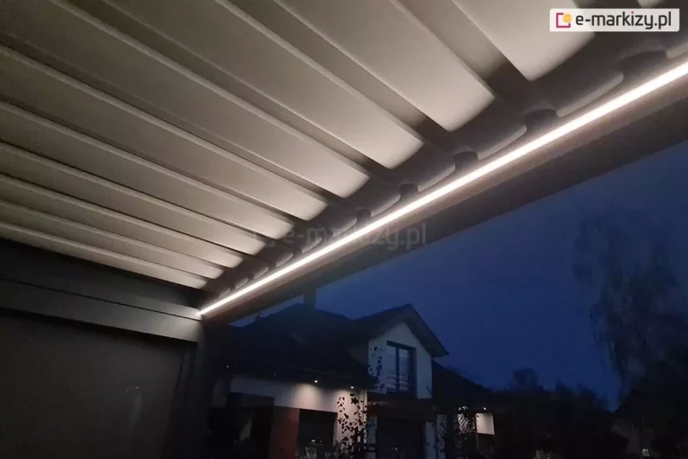 Zdjęcie przedstawia detal nowoczesnej pergoli tarasowej sfotografowanej po zmroku, z perspektywy dolnej, skupiającej się na systemie oświetlenia oraz spodniej stronie dachu. Głównym elementem kompozycji jest długa, liniowa listwa LED zamontowana wzdłuż wewnętrznej krawędzi ramy pergoli, która emituje jasne, ciepłe białe światło. Światło to efektownie oświetla krawędzie poziomo ułożonych lameli dachowych w kolorze jasnoszarym lub białym, podkreślając ich rytmiczny układ i profilowanie. Lamele są domknięte, tworząc zwartą płaszczyznę zadaszenia. W lewym dolnym rogu widoczny jest fragment ciemnej konstrukcji ramy oraz górna część opuszczonej rolety bocznej. W tle, pod zadaszeniem, wyłania się widok na nowoczesny dom jednorodzinny z jasną elewacją i licznymi przeszkleniami, w których odbija się wewnętrzne oświetlenie budynku. Całość otoczenia spowita jest w ciemnoniebieskiej, nocnej aurze, co tworzy silny kontrast z intensywnym pasem światła LED zintegrowanego z pergolą.
