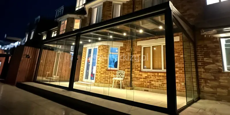 Zdjęcie prezentuje nocną scenerię tarasu, na którym zainstalowano nowoczesne, w pełni przeszklone zadaszenie Lunaro, zamontowane do klasycznej elewacji domu z czerwonej cegły. Konstrukcja w kolorze antracytowym jest wyposażona w system szklanych ścian przesuwnych, które tworzą zamknięty ogród zimowy, oświetlony od wewnątrz szeregiem jasnych, punktowych świateł LED zamontowanych w krokwiach dachowych. Przez transparentne szyby widoczne jest wnętrze tarasu z jasnymi drzwiami wejściowymi, białym krzesłem ażurowym oraz fragmentami murowanej ściany budynku. W tle widać bryłę piętrowego domu z podświetlonymi oknami oraz ciemny zarys drewnianego ogrodzenia posesji.