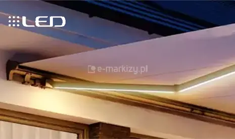 Wieczorne ujęcie prezentujące nowoczesne oświetlenie led do markiz. Podświetlenie ma formę liniowych listew zamontowanych wzdłuż dolnej krawędzi aluminiowych ramion, które emitują jasne, ciepłe lub zimne światło o barwie białej, skutecznie iluminując przestrzeń pod rozwiniętym poszyciem. Markiza o jasnym materiale jest zamontowana do elewacji budynku, a światło LED podkreśla geometrię konstrukcji oraz teksturę tkaniny. W lewym górnym rogu grafiki znajduje się biały napis LED z ikoną diod, sugerujący funkcjonalność systemu oświetleniowego. Całość tworzy nastrojową atmosferę na tarasie po zmroku, łącząc funkcję ochronną z dekoracyjną iluminacją strefy wypoczynkowej.