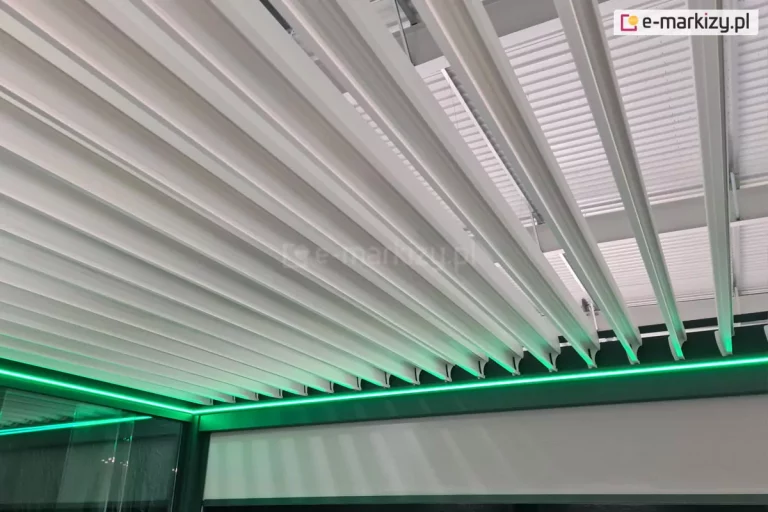 Zdjęcie prezentuje zbliżenie na system zadaszenia lamelowego nowoczesnej pergoli, wykonane pod kątem od dołu. Głównym elementem są białe, aluminiowe lamele, które znajdują się w pozycji otwartej (pionowej), co pozwala na wgląd w ich strukturę oraz profile mocujące. Wzdłuż wewnętrznej krawędzi ciemnoantracytowej ramy konstrukcyjnej zamontowano liniową taśmę LED, która emituje intensywne, neonowe światło o jaskrawozielonej barwie. Zielona poświata oświetla krawędzie lameli oraz górną część bocznej osłony typu screen, znajdującej się pod rynną. W lewym dolnym rogu widoczny jest fragment pionowego przeszklenia, w którym odbija się konstrukcja, sugerując, że system jest zamontowany w profesjonalnym salonie ekspozycyjnym. W tle, powyżej lameli, dostrzec można strukturę sufitu hali oraz elementy montażowe i uszczelniające systemu dachowego.