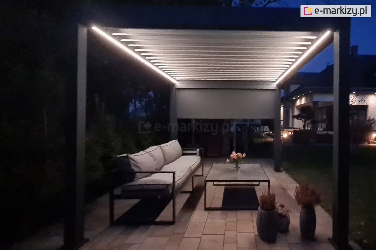 Zdjęcie przedstawia wolnostojącą pergolę bioklimatyczną o antracytowej konstrukcji, sfotografowaną w porze wieczornej z włączonym oświetleniem. Aluminiowa rama w kolorze ciemnego matu opiera się na czterech solidnych słupach, a jej zadaszenie stanowią białe, ruchome lamele, które są domknięte. Wzdłuż wewnętrznych krawędzi górnej ramy zamontowano liniowe oświetlenie LED, które emituje intensywne, ciepłe światło, rozjaśniając przestrzeń pod zadaszeniem i kontrastując z głębokim mrokiem otoczenia. Pod pergolą, na podłożu z jasnej kostki brukowej o beżowych odcieniach, urządzono elegancką strefę wypoczynkową. Znajduje się tam długa, nowoczesna sofa z jasnoszarymi poduszkami oraz prostokątny stolik kawowy z ciemnym stelażem, na którym ustawiono wazon z różowymi kwiatami. Obok mebli, w dolnej części kadru, widoczne są ozdobne donice z suchymi roślinami dekoracyjnymi. Na jednej z bocznych ścian konstrukcji zamontowano roletę zewnętrzną typu screen, która jest częściowo opuszczona. W tle, po prawej stronie, widać zarys bryły domu z podświetloną elewacją, co dodaje głębi nocnej scenerii ogrodu.