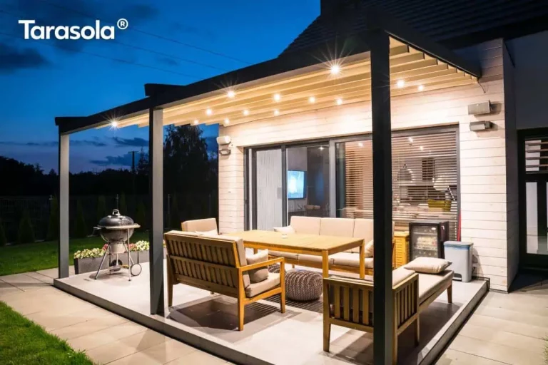 Zdjęcie prezentuje luksusową pergolę tarasową tarasola elegancy w aranżacji wieczornej, zamontowaną na nowoczesnym tarasie. Konstrukcja o grafitowym, aluminiowym stelażu jest zintegrowana z jasną elewacją budynku wyłożoną poziomymi deskami. Pod rozłożonym, jasnym zadaszeniem tkaninowym widoczne jest intensywne, punktowe oświetlenie LED, które tworzy ciepłą atmosferę i oświetla strefę wypoczynkową. Wyposażenie tarasu obejmuje komplet drewnianych mebli ogrodowych z jasnymi poduszkami, w tym sofę, fotele oraz stół, a także nowoczesny grill węglowy ustawiony z boku. Przeszklone drzwi przesuwne prowadzą do wnętrza domu, w którym widoczny jest salon. Całość instalacji tarasola elegancy podkreśla nowoczesny design budynku i funkcjonalność przestrzeni zewnętrznej po zmroku, otoczonej zadbanym trawnikiem i linią drzew w oddali.
