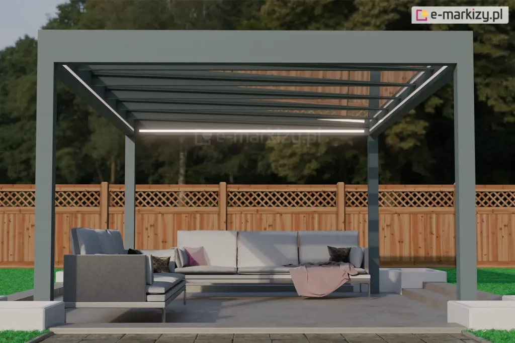 Nowoczesna pergola tarasowa Spekta Glass o konstrukcji wolnostojącej, umiejscowiona w ogrodzie na utwardzonym podłożu. Rama nośna oraz cztery słupy wsporcze zostały wykonane z profili aluminiowych w matowym, antracytowym kolorze, nadającym całości minimalistyczny wygląd. Zadaszenie składa się z równolegle rozmieszczonych profili, pomiędzy którymi znajdują się tafle szkła bezpiecznego, pod którymi zamontowano zintegrowane oświetlenie LED w formie liniowych pasów, emitujących ciepłe, białe światło. Pod konstrukcją zaaranżowano strefę wypoczynkową z nowoczesnym kompletem mebli ogrodowych, składającym się z dużej, narożnej sofy z jasnoszarymi poduszkami siedziskowymi i oparciowymi. Na sofie widoczne są dekoracyjne poduszki w kolorze ciemnym i fioletowym oraz różowy koc. Podłoże tarasu stanowią gładkie, jasnoszare płyty betonowe, które graniczą z pasem ciemniejszej kostki brukowej oraz zadbanym trawnikiem. W tle widoczne jest wysokie, drewniane ogrodzenie o konstrukcji panelowej z ozdobnym, kratkowanym zwieńczeniem, a za nim gęsta ściana zielonych drzew. Całość kompozycji przedstawia funkcjonalną i estetyczną przestrzeń rekreacyjną, idealnie wkomponowaną w architekturę nowoczesnego ogrodu.