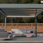 Nowoczesna pergola tarasowa Spekta Glass o konstrukcji wolnostojącej, umiejscowiona w ogrodzie na utwardzonym podłożu. Rama nośna oraz cztery słupy wsporcze zostały wykonane z profili aluminiowych w matowym, antracytowym kolorze, nadającym całości minimalistyczny wygląd. Zadaszenie składa się z równolegle rozmieszczonych profili, pomiędzy którymi znajdują się tafle szkła bezpiecznego, pod którymi zamontowano zintegrowane oświetlenie LED w formie liniowych pasów, emitujących ciepłe, białe światło. Pod konstrukcją zaaranżowano strefę wypoczynkową z nowoczesnym kompletem mebli ogrodowych, składającym się z dużej, narożnej sofy z jasnoszarymi poduszkami siedziskowymi i oparciowymi. Na sofie widoczne są dekoracyjne poduszki w kolorze ciemnym i fioletowym oraz różowy koc. Podłoże tarasu stanowią gładkie, jasnoszare płyty betonowe, które graniczą z pasem ciemniejszej kostki brukowej oraz zadbanym trawnikiem. W tle widoczne jest wysokie, drewniane ogrodzenie o konstrukcji panelowej z ozdobnym, kratkowanym zwieńczeniem, a za nim gęsta ściana zielonych drzew. Całość kompozycji przedstawia funkcjonalną i estetyczną przestrzeń rekreacyjną, idealnie wkomponowaną w architekturę nowoczesnego ogrodu.