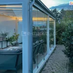 Widok na nowoczesny ogród zimowy wykonany przylegający do budynku mieszkalnego. Konstrukcja posiada bezramowe, przesuwne panele szklane Miros, zamontowane w białych, aluminiowych profilach. We wnętrzu znajduje się strefa wypoczynkowa z dwiema ciemnoszarymi sofami ogrodowymi ustawionymi wokół prostokątnego stołu z jasnym blatem, na którym leżą kubki, dokumenty i niewielkie przedmioty. Przy lewej ścianie ustawiony jest długi czarny regał z kilkoma doniczkami z zielonymi roślinami ozdobnymi. Wewnątrz ogrodu zimowego zamontowane są panele sufitowe z podświetleniem LED o ciepłej barwie. Na zewnątrz, po prawej stronie, widać brukowaną ścieżkę otoczoną bujną roślinnością, w tym ciemnozielone krzewy. W tle widoczny fragment ogrodu oraz dachy sąsiednich domów pod wieczornym niebem.