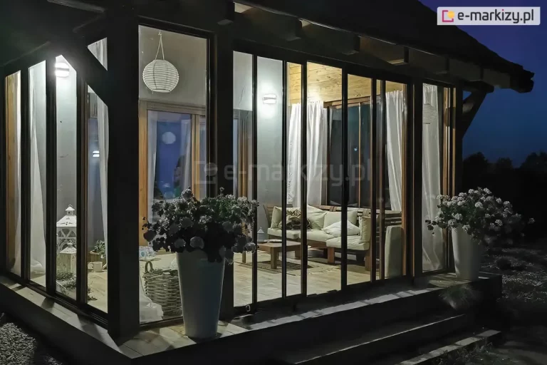 zabudowany taras w porze nocnej, intensywnie oświetlony od wewnątrz ciepłym światłem. Konstrukcja oparta na ciemnych profilach posiada system wysokich drzwi przesuwnych oraz stałych przeszkleń bocznych, które sięgają od podłogi aż po konstrukcję dachu. Wewnątrz przytulnej przestrzeni widoczne są meble wypoczynkowe z jasnymi poduszkami, stolik kawowy oraz białe lampiony dekoracyjne i wisząca lampa w kształcie kuli. Na zewnątrz, przed szklaną ścianą, ustawiono duże, białe donice z roślinami kwitnącymi, które flankują wejście. Drewniana podłoga tarasu oraz jasne zasłony wewnątrz podkreślają domowy charakter ogrodu zimowego. Całość kompozycji kontrastuje z ciemnym, nocnym niebem widocznym w tle.