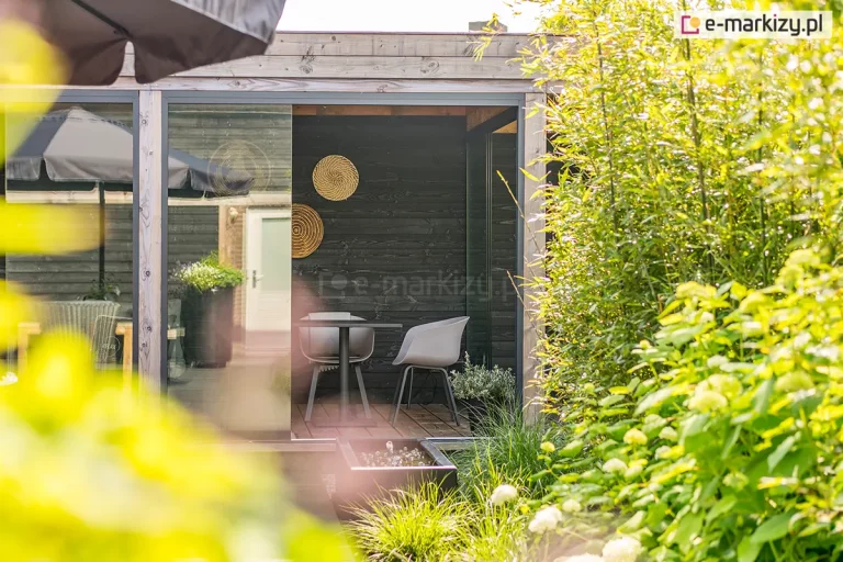 Szerokie ujęcie na otwartą przestrzeń nowoczesnego ogrodu letniego z wykorzystaniem systemu przesuwnych ścian szklanych Auris. Zdjęcie przedstawia kaskadowo zsunięte panele z przezroczystego szkła hartowanego, które tworzą szerokie światło przejścia, łącząc wnętrze altany z tarasem. Wnętrze charakteryzuje się czarnymi ścianami z drewna i minimalistycznym zestawem jadalnianym (czarny stolik i nowoczesne, szare krzesła). Na zewnątrz widoczna jest bujna zieleń ogrodowa, w tym gęste krzewy i trawy ozdobne, które częściowo przysłaniają kadr, nadając mu naturalny charakter. System Auris w kolorze antracytowym idealnie komponuje się z nowoczesną architekturą ogrodową, oferując elastyczność w zarządzaniu przestrzenią w zależności od pogody.