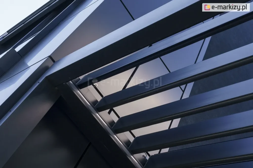 Pergola lamelowa SB400 w kolorze antracytowym zamocowana przy ścianie budynku o elewacji wykonanej w tym samym kolorze. Przez otwarte lamele pod pergolę wpada światło, a między poszczególnymi lamelami można zauważyć cięgno mechanizmu otwierania dachu.
