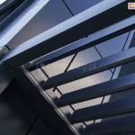 Pergola lamelowa SB400 w kolorze antracytowym zamocowana przy ścianie budynku o elewacji wykonanej w tym samym kolorze. Przez otwarte lamele pod pergolę wpada światło, a między poszczególnymi lamelami można zauważyć cięgno mechanizmu otwierania dachu.