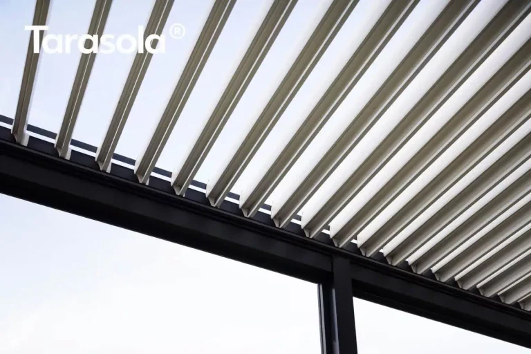 Zbliżenie na detal konstrukcyjny dachu nowoczesnej pergoli Essential Tarasola, prezentujące mechanizm ruchomych lameli. Zdjęcie wykonane z perspektywy dolnej ukazuje rzędy równolegle ułożonych, aluminiowych lameli w kolorze białym, które są w tej chwili częściowo otwarte (uchylone pod kątem). Taka konfiguracja modelu Essential Tarasola pozwala na precyzyjną regulację ilości wpadającego światła słonecznego oraz zapewnia naturalną cyrkulację powietrza pod zadaszeniem. Lamele mają charakterystyczny, profilowany kształt z wyraźnymi krawędziami, co wspomaga odprowadzanie wody deszczowej do systemu rynnowego. Konstrukcja wsporcza dachu, widoczna w dolnej części kadru, wykonana jest z solidnych profili aluminiowych malowanych proszkowo na kolor ciemnoantracytowy, co tworzy kontrast z jasnym wypełnieniem zadaszenia. Widoczne jest miejsce styku poziomej belki ramy z pionowym słupem nośnym pergoli Essential Tarasola, co podkreśla precyzję łączeń i wysoką jakość wykończenia produktu. W tle, pomiędzy uchylonymi lamelami, przebija jasne, bezchmurne niebo.