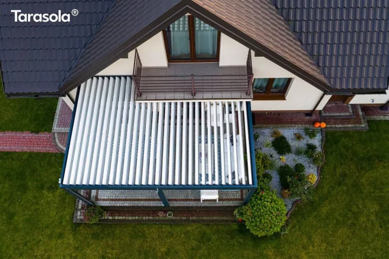 Widok z lotu ptaka przedstawia nowoczesną pergolę tarasową Essential Tarasola zamontowaną przy ścianie piętrowego domu jednorodzinnego. Konstrukcja zadaszenia opiera się na ciemnoszarym, antracytowym stelażu wykonanym z metalowych profili, który przylega bezpośrednio do elewacji budynku pod balkonem znajdującym się na wyższej kondygnacji. Dach pergoli składa się z białych, równolegle ułożonych lameli, które są w tej chwili częściowo otwarte, tworząc pasy światła i cienia na powierzchni tarasu pod nimi. Pod zadaszeniem widoczna jest przestrzeń wypoczynkowa z jasnymi meblami ogrodowymi ustawionymi na nawierzchni z szarej kostki brukowej, która jest wykończona ozdobną obwódką z ciemniejszej kostki. Dom posiada białą elewację oraz dach pokryty ciemną dachówką o falistym profilu. Nad pergolą znajduje się balkon z metalową, minimalistyczną balustradą w kolorze brązowym oraz przeszklone drzwi balkonowe osadzone w drewnianej stolarce. Otoczenie budynku to zadbany, zielony trawnik, przez który przebiega ścieżka z czerwonej kostki brukowej prowadząca do tarasu. Po prawej stronie pergoli znajduje się ozdobna rabata ogrodowa wysypana drobnym, jasnym żwirem, na której posadzono różnorodne rośliny ozdobne, w tym niskie krzewy iglaste i liściaste oraz większy, gęsty krzew przy samym narożniku konstrukcji. Na krawędzi opaski wokół domu, niedaleko wejścia, widoczne są dwie pomarańczowe dynie stanowiące element dekoracyjny. Całość kompozycji ukazuje funkcjonalne połączenie nowoczesnej architektury ogrodowej z klasyczną bryłą domu.