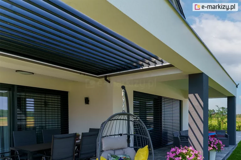 Nowoczesny budynek mieszkalny z tarasem wyposażonym w dach lamelowy SB400R Selt. Aluminiowe lamele dachowe w kolorze antracytowym są ustawione w pozycji otwartej (pionowej), co zapewnia maksymalny dopływ światła dziennego i pełną wentylację strefy wypoczynkowej. Rama zadaszenia posiada zintegrowane oświetlenie liniowe LED, które jest widoczne wzdłuż wewnętrznych krawędzi konstrukcji. Pod pergolą znajduje się komplet mebli tarasowych: ciemny stół z krzesłami oraz wiszący fotel ogrodowy z szarymi poduszkami. Jasna elewacja domu kontrastuje z ciemnymi słupami nośnymi pergoli oraz antracytowymi żaluzjami fasadowymi zamontowanymi w oknach. Na tarasie wyłożonym płytami betonowymi ustawiono donice z fioletowymi kwiatami, a w tle widoczna jest otwarta przestrzeń z zieloną roślinnością pod lekko zachmurzonym niebem.