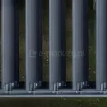 Zbliżenie z góry na mechanizm otwierany dachu pergoli tarasowej Gloria, prezentujące system aluminiowych lameli w kolorze antracytowym. Lamele są ustawione w pozycji pionowej (całkowicie otwartej), co odsłania widok na podłoże znajdujące się pod konstrukcją. W dolnej części kadru widoczna jest szyna prowadząca oraz precyzyjne elementy montażowe i przeguby, które odpowiadają za synchroniczny obrót każdego profilu dachowego. Detal techniczny podkreśla matowe wykończenie aluminium oraz powtarzalność i symetrię elementów mechanicznych dachu bioklimatycznego.