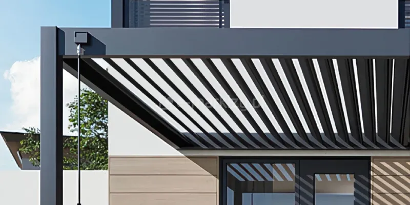 Zbliżenie na fragment konstrukcji nowoczesnej pergoli tarasowej Rubio Soft, wykonanej z aluminium w kolorze antracytowym, skupiające się na mechanizmie sterowania zadaszeniem. Zdjęcie prezentuje otwarte lamele dachowe ustawione pod kątem, co tworzy rytmiczny wzór ukośnych, równoległych pasów, przez które prześwituje jasne niebo. Na przedniej, poziomej belce ramy zamontowana jest czarna, kwadratowa puszka mechanizmu, z której wyprowadzona jest długa, pionowa korba sterująca o matowym wykończeniu. W tle widoczna jest jasna elewacja budynku z fragmentem okna oraz pionowe pasy cienia rzucane przez otwarte lamele na ścianę wykończoną drewnopodobnymi panelami. Konstrukcja charakteryzuje się ostrymi krawędziami i minimalistycznym wykończeniem, typowym dla nowoczesnej architektury ogrodowej. Perspektywa ujęcia od dołu podkreśla przestrzenną strukturę dachu oraz precyzję wykonania elementów ruchomych.