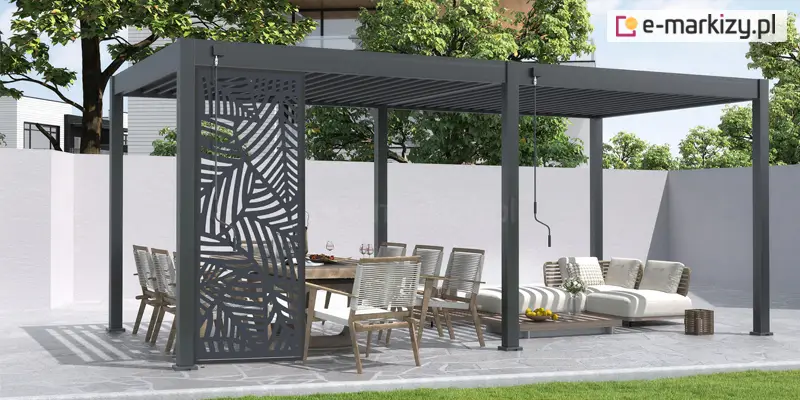Wizualizacja nowoczesnej, wolnostojącej pergoli tarasowej o podwójnym module w kolorze antracytowym, usytuowanej na nowoczesnym patio wyłożonym jasnym kamieniem. Konstrukcja z płaskim, lamelowym dachem posiada środkowy słup wsporczy, przy którym zamontowano pionową korbę manualną do regulacji zadaszenia. Po lewej stronie frontowej części pergoli zainstalowano ozdobny panel ażurowy z wyciętym motywem dużych liści palmowych, który pełni funkcję dekoracyjną i przesłaniającą. Pod pergolą zaaranżowano dwie strefy: jadalnianą z dużym drewnianym stołem i nowoczesnymi krzesłami o jasnych siedziskach oraz wypoczynkową z białymi, modułowymi meblami ogrodowymi i niskim stolikiem kawowym. W tle widoczny jest minimalistyczny, biały mur, za którym wyrastają korony zielonych drzew. Całość kompozycji dopełnia fragment soczyście zielonego trawnika na pierwszym planie, a światło słoneczne rzuca wyraźne, geometryczne cienie konstrukcji na podłoże.
