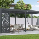 Antracytowa wolnostojąca pergola lamelowa Marlo o modułowej konstrukcji, zamontowana na tarasie wyłożonym nieregularnymi płytami kamiennymi. Cechą charakterystyczną tej aranżacji jest ozdobny panel narożny z wyciętym laserowo wzorem liści palmowych, zamontowany na jednym z przęseł bocznych, który pełni funkcję dekoracyjną i zapewnia częściową prywatność. Zadaszenie składa się z dwóch sekcji lamel, które są ustawione w pozycji zamkniętej. Na konstrukcji widoczne są dwa mechanizmy korbowe do ręcznego sterowania dachem. Pod pergolą znajduje się strefa jadalniana z drewnianym stołem i krzesłami o białych plecionkach oraz strefa wypoczynkowa z niskimi, jasnymi sofami modułowymi ustawionymi na drewnianym podeście. Całość otoczona jest białym murkiem, za którym widoczne są drzewa liściaste oraz fragment nowoczesnej elewacji budynku. Na pierwszym planie znajduje się równo przystrzyżony zielony trawnik.