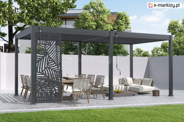 Antracytowa wolnostojąca pergola lamelowa Marlo o modułowej konstrukcji, zamontowana na tarasie wyłożonym nieregularnymi płytami kamiennymi. Cechą charakterystyczną tej aranżacji jest ozdobny panel narożny z wyciętym laserowo wzorem liści palmowych, zamontowany na jednym z przęseł bocznych, który pełni funkcję dekoracyjną i zapewnia częściową prywatność. Zadaszenie składa się z dwóch sekcji lamel, które są ustawione w pozycji zamkniętej. Na konstrukcji widoczne są dwa mechanizmy korbowe do ręcznego sterowania dachem. Pod pergolą znajduje się strefa jadalniana z drewnianym stołem i krzesłami o białych plecionkach oraz strefa wypoczynkowa z niskimi, jasnymi sofami modułowymi ustawionymi na drewnianym podeście. Całość otoczona jest białym murkiem, za którym widoczne są drzewa liściaste oraz fragment nowoczesnej elewacji budynku. Na pierwszym planie znajduje się równo przystrzyżony zielony trawnik.