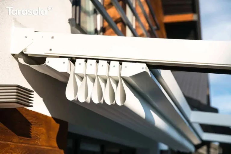 Zbliżenie na techniczny detal pakietu tkaniny rzymskiej białej pergoli tarasowej Tarasola Simple w miejscu jej mocowania do elewacji budynku. Widoczny jest precyzyjny mechanizm składania materiału, gdzie jasna tkanina techniczna układa się w równe, pionowe fałdy podwieszone pod aluminiową szyną prowadzącą. Konstrukcja mocująca oraz profile poprzeczne są lakierowane proszkowo na kolor biały i połączone za pomocą stalowych śrub. W tle widać fragment ściany z białym tynkiem, ozdobne panele elewacyjne o strukturze ciemnego drewna oraz nowoczesną oprawę oświetleniową o geometrycznym kształcie. Perspektywa z dołu podkreśla stabilność aluminiowego stelaża oraz estetykę wykończenia ruchomego zadaszenia.