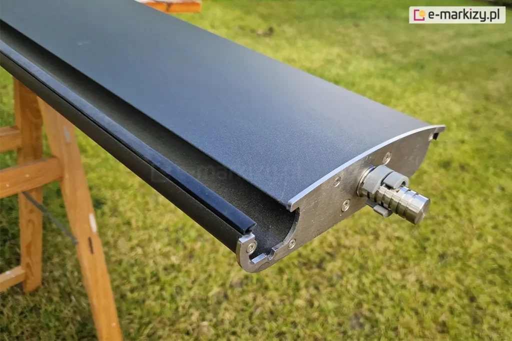 zbliżenie na pojedynczą lamelę dachu pergoli Selt SB400, przygotowaną do montażu. Aluminiowy profil o aerodynamicznym kształcie jest pomalowany proszkowo na matowy kolor antracytowy. Na bocznej, srebrnej krawędzi (deklu) widoczny jest wystający metalowy trzpień z pierścieniami prowadzącymi, który stanowi element obrotowy montowany w listwach bocznych konstrukcji. Wzdłuż dolnej krawędzi lameli zamocowana jest czarna, gumowa uszczelka, zapewniająca szczelność dachu po jego zamknięciu. Element spoczywa na drewnianym stojaku montażowym (kobyłce), a w tle widoczny jest zielony trawnik. Perspektywa pozwala na dokładne przyjrzenie się budowie wewnętrznej profilu oraz precyzji wykończenia detali mechanicznych.
