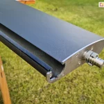 zbliżenie na pojedynczą lamelę dachu pergoli Selt SB400, przygotowaną do montażu. Aluminiowy profil o aerodynamicznym kształcie jest pomalowany proszkowo na matowy kolor antracytowy. Na bocznej, srebrnej krawędzi (deklu) widoczny jest wystający metalowy trzpień z pierścieniami prowadzącymi, który stanowi element obrotowy montowany w listwach bocznych konstrukcji. Wzdłuż dolnej krawędzi lameli zamocowana jest czarna, gumowa uszczelka, zapewniająca szczelność dachu po jego zamknięciu. Element spoczywa na drewnianym stojaku montażowym (kobyłce), a w tle widoczny jest zielony trawnik. Perspektywa pozwala na dokładne przyjrzenie się budowie wewnętrznej profilu oraz precyzji wykończenia detali mechanicznych.