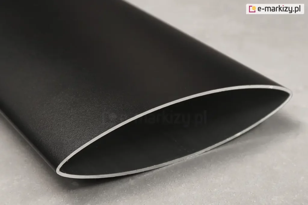 Zbliżenie na pojedynczy profil aluminiowy stanowiący element panelu żaluzjowego Louvre, prezentujący jego techniczny przekrój. Element ma charakterystyczny, aerodynamiczny kształt soczewki lub wydłużonej elipsy o ostrych krawędziach bocznych. Powierzchnia profilu jest wykończona matową, lekko ziarnistą strukturą w kolorze głębokiego antracytu lub czerni. Na krawędzi cięcia widoczna jest naturalna, srebrzysta barwa aluminium, co podkreśla grubość ścianki profilu. Przedmiot spoczywa na jasnoszarym, jednolitym podłożu o delikatnie porowatej fakturze, przypominającej beton lub kamień. Oświetlenie jest miękkie i rozproszone, co pozwala na dokładne zaobserwowanie satynowego połysku na obłej powierzchni detalu oraz precyzji jego wykonania.