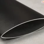 Zbliżenie na pojedynczy profil aluminiowy stanowiący element panelu żaluzjowego Louvre, prezentujący jego techniczny przekrój. Element ma charakterystyczny, aerodynamiczny kształt soczewki lub wydłużonej elipsy o ostrych krawędziach bocznych. Powierzchnia profilu jest wykończona matową, lekko ziarnistą strukturą w kolorze głębokiego antracytu lub czerni. Na krawędzi cięcia widoczna jest naturalna, srebrzysta barwa aluminium, co podkreśla grubość ścianki profilu. Przedmiot spoczywa na jasnoszarym, jednolitym podłożu o delikatnie porowatej fakturze, przypominającej beton lub kamień. Oświetlenie jest miękkie i rozproszone, co pozwala na dokładne zaobserwowanie satynowego połysku na obłej powierzchni detalu oraz precyzji jego wykonania.