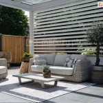 Zdjęcie przedstawia aranżację nowoczesnego tarasu z wykorzystaniem białego panelu żaluzjowego Louvre jako bocznej osłony pergoli. Białe, poziome lamele o eliptycznym przekroju są zamontowane na stałe w pionowych profilach, tworząc ażurową ścianę, która zapewnia prywatność, jednocześnie przepuszczając światło i powietrze. Na pierwszym planie znajduje się strefa wypoczynkowa z meblami wykonanymi z technorattanu o grubym splocie w kolorze szarym, obejmująca sofę z jasnymi poduszkami oraz pasujący fotel. Pomiędzy meblami ułożono jasny dywan zewnętrzny z delikatnym wzorem, na którym stoi niski, nowoczesny stolik kawowy w kolorze jasnoszarym. Dekorację dopełniają rośliny w donicach, w tym niewielkie drzewko oliwne, oraz drewniane elementy ozdobne w formie pni. W tle, za panelem, widoczne jest drewniane ogrodzenie oraz gęsta zielona roślinność ogrodowa, co podkreśla przytulny i elegancki charakter przestrzeni.