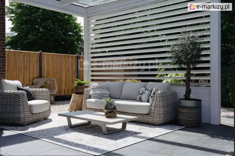 Zdjęcie przedstawia aranżację nowoczesnego tarasu z wykorzystaniem białego panelu żaluzjowego Louvre jako bocznej osłony pergoli. Białe, poziome lamele o eliptycznym przekroju są zamontowane na stałe w pionowych profilach, tworząc ażurową ścianę, która zapewnia prywatność, jednocześnie przepuszczając światło i powietrze. Na pierwszym planie znajduje się strefa wypoczynkowa z meblami wykonanymi z technorattanu o grubym splocie w kolorze szarym, obejmująca sofę z jasnymi poduszkami oraz pasujący fotel. Pomiędzy meblami ułożono jasny dywan zewnętrzny z delikatnym wzorem, na którym stoi niski, nowoczesny stolik kawowy w kolorze jasnoszarym. Dekorację dopełniają rośliny w donicach, w tym niewielkie drzewko oliwne, oraz drewniane elementy ozdobne w formie pni. W tle, za panelem, widoczne jest drewniane ogrodzenie oraz gęsta zielona roślinność ogrodowa, co podkreśla przytulny i elegancki charakter przestrzeni.