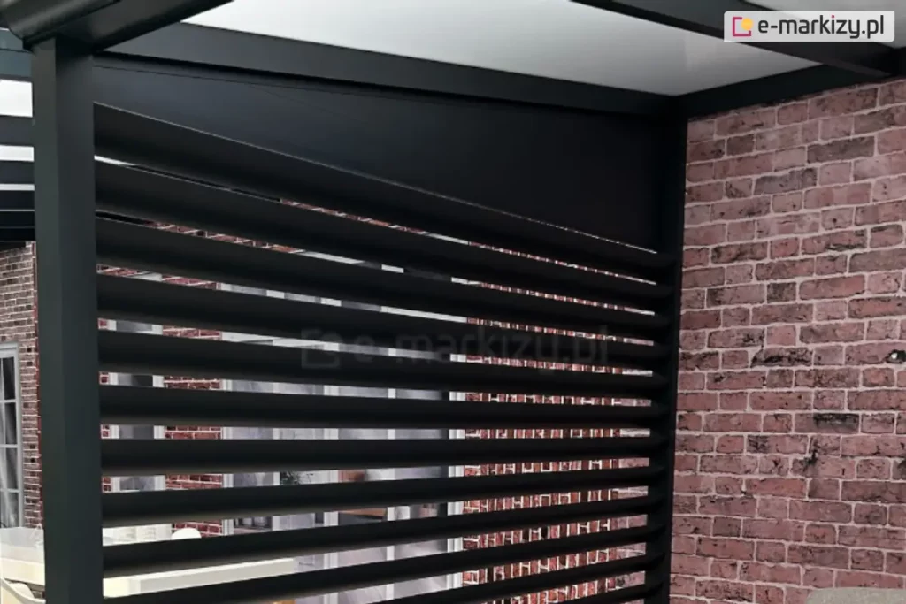 Zdjęcie prezentuje aluminiowy panel żaluzjowy louvre zamontowany jako boczne wypełnienie nowoczesnej pergoli, widziany od strony wewnętrznej tarasu. Konstrukcja składa się z szeregu równoległych, poziomych lameli o eliptycznym przekroju w kolorze antracytowym, które są osadzone w pionowej ramie. Nad żaluzjami znajduje się pełny, trójkątny panel maskujący, który estetycznie wypełnia przestrzeń pod skośnym dachem zadaszenia. Po prawej stronie widoczny jest pionowy słup konstrukcyjny pergoli przylegający do ściany budynku wykonanej z dekoracyjnej, czerwonej cegły. Przez prześwity między lamelami widać fragmenty wnętrza oraz przeszklenia, co obrazuje funkcję osłony jako elementu zapewniającego prywatność przy zachowaniu dopływu naturalnego światła.