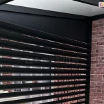 Zdjęcie prezentuje aluminiowy panel żaluzjowy louvre zamontowany jako boczne wypełnienie nowoczesnej pergoli, widziany od strony wewnętrznej tarasu. Konstrukcja składa się z szeregu równoległych, poziomych lameli o eliptycznym przekroju w kolorze antracytowym, które są osadzone w pionowej ramie. Nad żaluzjami znajduje się pełny, trójkątny panel maskujący, który estetycznie wypełnia przestrzeń pod skośnym dachem zadaszenia. Po prawej stronie widoczny jest pionowy słup konstrukcyjny pergoli przylegający do ściany budynku wykonanej z dekoracyjnej, czerwonej cegły. Przez prześwity między lamelami widać fragmenty wnętrza oraz przeszklenia, co obrazuje funkcję osłony jako elementu zapewniającego prywatność przy zachowaniu dopływu naturalnego światła.