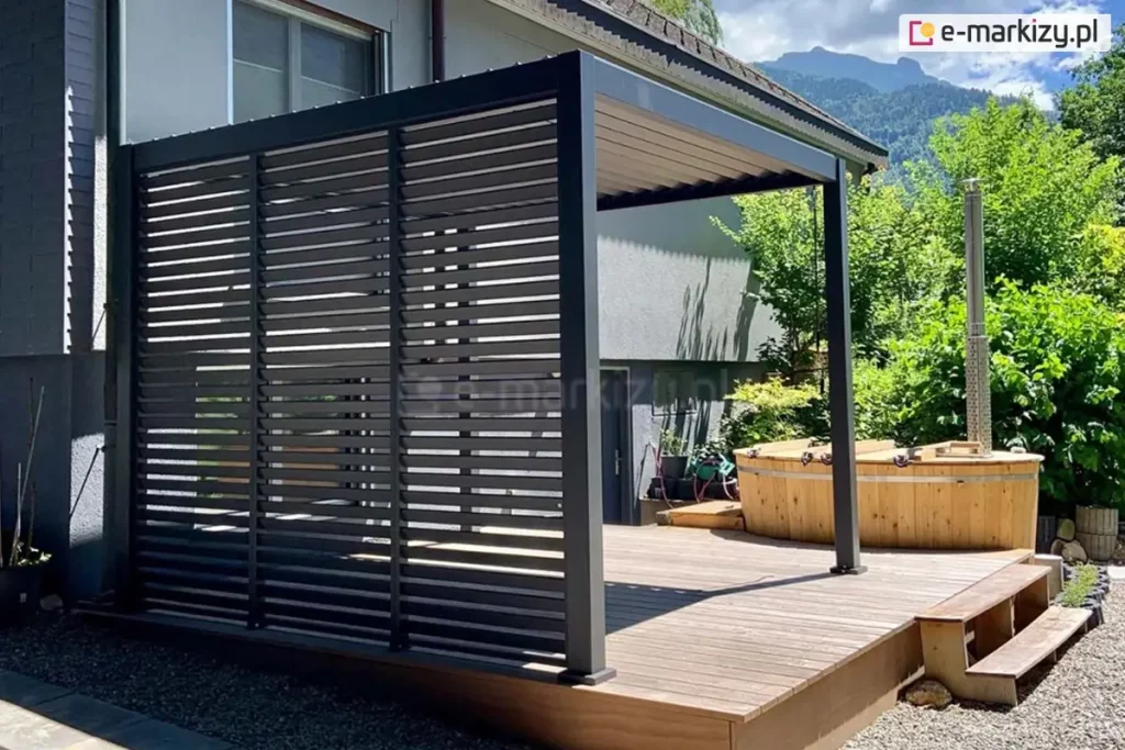 Zdjęcie prezentuje nowoczesną pergolę aluminiową Gloria w kolorze antracytowym, zainstalowaną na drewnianym tarasie przy budynku mieszkalnym. Głównym elementem zabudowy są boczne panele żaluzjowe, które składają się z trzech pionowych sekcji wypełnionych stałymi, poziomymi lamelami. Konstrukcja ta tworzy szczelną barierę wizualną, zapewniając prywatność i ochronę przed słońcem, jednocześnie nadając bryle nowoczesny, geometryczny wygląd. Zadaszenie pergoli wykonane jest z ruchomych lameli dachowych, które są widoczne od spodu w pozycji lekko uchylonej. Obok pergoli na tarasie znajduje się okrągła, drewniana balia ogrodowa (hot tub) z metalowym piecem zewnętrznym i kominem. Całość kompozycji dopełnia widok na zielony ogród, wysokie drzewa iglaste oraz malownicze zbocze góry w tle pod lekko zachmurzonym niebem.