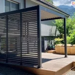 Zdjęcie prezentuje nowoczesną pergolę aluminiową Gloria w kolorze antracytowym, zainstalowaną na drewnianym tarasie przy budynku mieszkalnym. Głównym elementem zabudowy są boczne panele żaluzjowe, które składają się z trzech pionowych sekcji wypełnionych stałymi, poziomymi lamelami. Konstrukcja ta tworzy szczelną barierę wizualną, zapewniając prywatność i ochronę przed słońcem, jednocześnie nadając bryle nowoczesny, geometryczny wygląd. Zadaszenie pergoli wykonane jest z ruchomych lameli dachowych, które są widoczne od spodu w pozycji lekko uchylonej. Obok pergoli na tarasie znajduje się okrągła, drewniana balia ogrodowa (hot tub) z metalowym piecem zewnętrznym i kominem. Całość kompozycji dopełnia widok na zielony ogród, wysokie drzewa iglaste oraz malownicze zbocze góry w tle pod lekko zachmurzonym niebem.