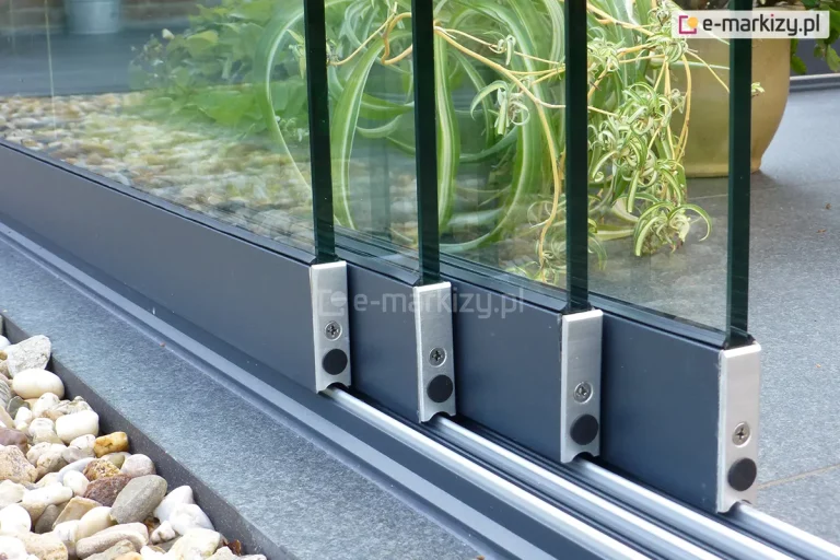 Zbliżenie na dolne profile prowadzące (wózki jezdne) bezramowego systemu szklanego Auris. Zdjęcie prezentuje cztery tafle szkła hartowanego osadzone w eleganckich, antracytowych profilach dolnych, zakończonych srebrnymi elementami konstrukcyjnymi z widocznymi śrubami montażowymi i czarnymi zaślepkami. System porusza się po wielotorowej szynie dolnej, która jest zamontowana na styku ciemnoszarych płyt tarasowych oraz ozdobnego pasa z jasnych kamieni rzecznych (otoczaków). W tle, za przezroczystym przeszkleniem, widoczne są zielone rośliny doniczkowe, co podkreśla minimalistyczny design i wysoką jakość wykończenia detali technicznych zabudowy.