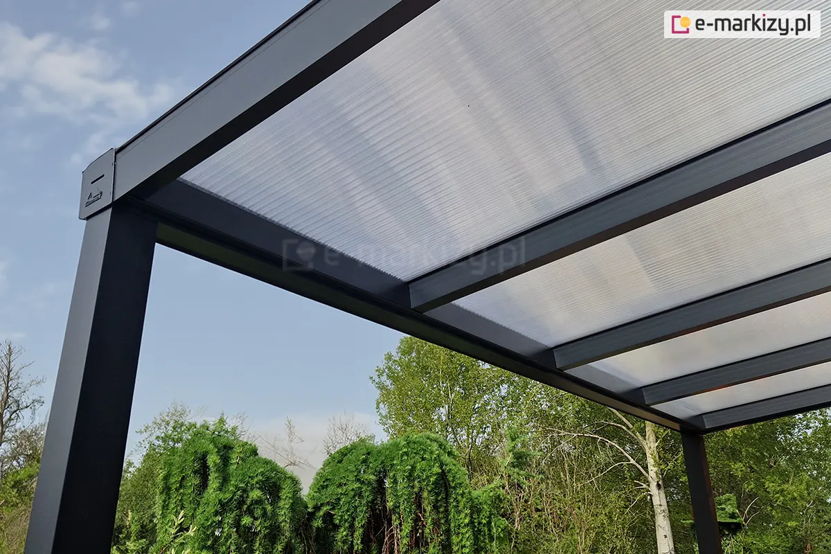 Widok na panele poliwęglanowe zamocowane w krokwiach zadaszenia tarasu od spodu. W tle za zadaszeniem widoczne drzewa z zielonymi liśćmi oraz błękitne niebo.