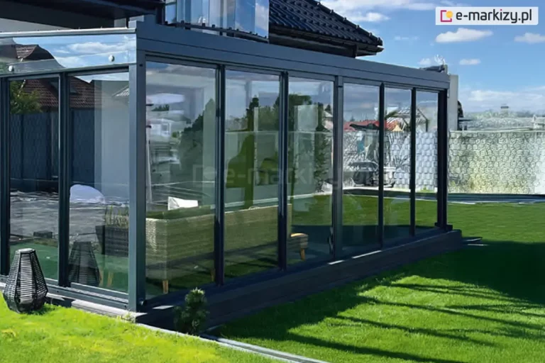 Luksusowa zabudowa tarasowa przylegającą do nowoczesnego domu jednorodzinnego z ciemnym dachem. Konstrukcja w kolorze antracytowym posiada w pełni przeszklone ściany wykonane z wielu wąskich paneli przesuwnych w systemie ramowym Sotro. Wewnątrz widać nowoczesny narożnik technorattanowy w kolorze beżowym. Zabudowa stoi na ciemnej, systemowej podbudowie, która jest zlicowana z poziomem trawnika. Duże tafle szkła odbijają okoliczne budynki oraz błękitne niebo z białymi chmurami. W tle po prawej stronie widoczne jest wysokie, dekoracyjne ogrodzenie.