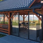 Drewniana konstrukcja restauracyjnego tarasu lub pawilonu z dachem pokrytym czarną dachówką, w którą wkomponowano system przesuwnych paneli szklanych. Szkielet wykonany jest z masywnych belek drewnianych w ciepłym odcieniu brązu, a przeszklenia osadzone są w czarnych, dyskretnych ramkach aluminiowych. Panele szklane pełnią funkcję drzwi wejściowych oraz bocznych ścianek osłonowych, chroniąc wnętrze przed wiatrem bez ograniczania widoczności na górzysty krajobraz. Wewnątrz obiektu widoczne są czarne stoliki i krzesła przygotowane dla gości, a na jednej z belek umieszczono tabliczkę z napisem informacyjnym. Przed wejściem znajduje się chodnik z szarej kostki brukowej, a po prawej stronie, tuż obok czarnej rynny odpływowej, na elewacji zamontowano nowoczesną, prostokątną lampę zewnętrzną.