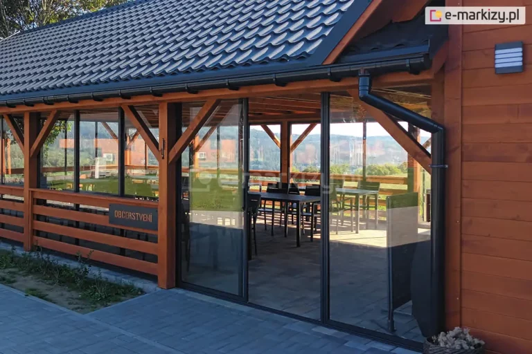 Drewniana konstrukcja restauracyjnego tarasu lub pawilonu z dachem pokrytym czarną dachówką, w którą wkomponowano system przesuwnych paneli szklanych. Szkielet wykonany jest z masywnych belek drewnianych w ciepłym odcieniu brązu, a przeszklenia osadzone są w czarnych, dyskretnych ramkach aluminiowych. Panele szklane pełnią funkcję drzwi wejściowych oraz bocznych ścianek osłonowych, chroniąc wnętrze przed wiatrem bez ograniczania widoczności na górzysty krajobraz. Wewnątrz obiektu widoczne są czarne stoliki i krzesła przygotowane dla gości, a na jednej z belek umieszczono tabliczkę z napisem informacyjnym. Przed wejściem znajduje się chodnik z szarej kostki brukowej, a po prawej stronie, tuż obok czarnej rynny odpływowej, na elewacji zamontowano nowoczesną, prostokątną lampę zewnętrzną.