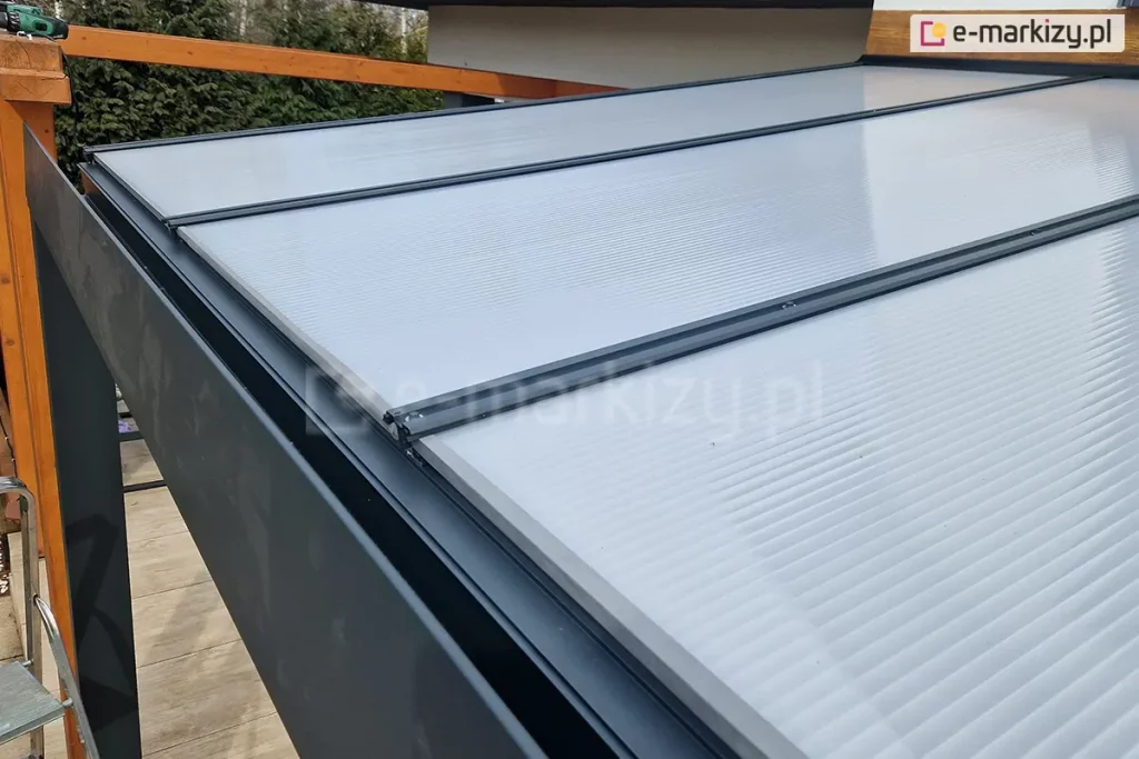 Zadaszenie tarasowe z aluminium w kolorze antracytowym z pokryciem z płyt poliwęglanowych w kolorze mlecznym. Specjalne połączenia płyt z konstrukcją zabezpieczają powierzchnię zadaszenia przed warunkami atmosferycznymi oraz przed zanieczyszczeniem poliwęglanu. W tle widoczna drewniana konstrukcja z pozostawionym na niej elektronarzędziem.
