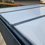 Zadaszenie tarasowe z aluminium w kolorze antracytowym z pokryciem z płyt poliwęglanowych w kolorze mlecznym. Specjalne połączenia płyt z konstrukcją zabezpieczają powierzchnię zadaszenia przed warunkami atmosferycznymi oraz przed zanieczyszczeniem poliwęglanu. W tle widoczna drewniana konstrukcja z pozostawionym na niej elektronarzędziem.