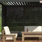 Widok na wnętrze nowoczesnej pergoli aluminiowej Gloria w kolorze antracytowym, wyposażonej w system ruchomych paneli żaluzjowych. Ściana boczna składa się z dwóch modułów wypełnionych poziomymi, czarnymi lamelami, które są ustawione w pozycji otwartej, tworząc ażurową przesłonę. Taka konfiguracja zapewnia skuteczną ochronę przed bezpośrednim nasłonecznieniem przy zachowaniu pełnej cyrkulacji powietrza i widoku na ogród. Zadaszenie konstrukcji również stanowią ruchome lamele dachowe, ustawione pod kątem, co pozwala na precyzyjną regulację ilości światła wpadającego do wnętrza. W strefie wypoczynkowej ustawiono drewniane meble ogrodowe: szeroki fotel z jasnymi poduszkami oraz niski stolik kawowy. Na pierwszym planie, przy lewym słupie nośnym, widoczna jest korba służąca do ręcznego sterowania mechanizmem obrotu lameli. W tle widać nasłoneczniony ogród z bogatą roślinnością i drzewami, co podkreśla relaksacyjny charakter przestrzeni tarasowej.