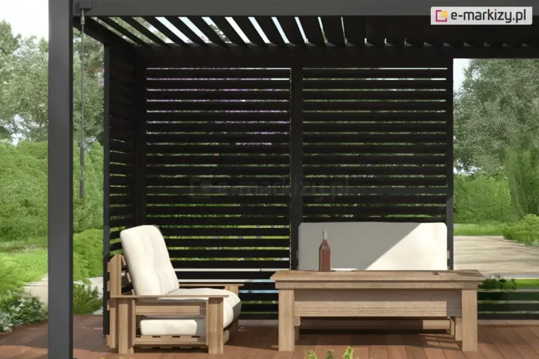 Widok na wnętrze nowoczesnej pergoli aluminiowej Gloria w kolorze antracytowym, wyposażonej w system ruchomych paneli żaluzjowych. Ściana boczna składa się z dwóch modułów wypełnionych poziomymi, czarnymi lamelami, które są ustawione w pozycji otwartej, tworząc ażurową przesłonę. Taka konfiguracja zapewnia skuteczną ochronę przed bezpośrednim nasłonecznieniem przy zachowaniu pełnej cyrkulacji powietrza i widoku na ogród. Zadaszenie konstrukcji również stanowią ruchome lamele dachowe, ustawione pod kątem, co pozwala na precyzyjną regulację ilości światła wpadającego do wnętrza. W strefie wypoczynkowej ustawiono drewniane meble ogrodowe: szeroki fotel z jasnymi poduszkami oraz niski stolik kawowy. Na pierwszym planie, przy lewym słupie nośnym, widoczna jest korba służąca do ręcznego sterowania mechanizmem obrotu lameli. W tle widać nasłoneczniony ogród z bogatą roślinnością i drzewami, co podkreśla relaksacyjny charakter przestrzeni tarasowej.