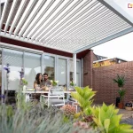 Zdjęcie przedstawia nowoczesną białą pergolę lamelową Gloria zamontowaną na tarasie przy budynku mieszkalnym. Konstrukcja pergoli opiera się na grubych, kwadratowych profilach aluminiowych w kolorze białym, a jej zadaszenie stanowią ruchome, równolegle ułożone lamele, które są w tym momencie częściowo otwarte, przepuszczając rozproszone światło słoneczne. Pergola jest przystawiona jedną stroną do elewacji budynku o ciemnoczerwonym wykończeniu nad oknami tarasowymi, a całość wspiera się na pionowych słupach zakotwiczonych w podłożu. Pod zadaszeniem znajduje się zestaw mebli ogrodowych składający się z białego stołu oraz dopasowanych krzeseł z szarymi poduszkami, przy których siedzi para osób w trakcie posiłku. Taras wyłożony jest deskami kompozytowymi lub drewnianymi w naturalnym kolorze, a w tle widać ceglany mur oraz fragmenty sąsiedniej zabudowy z dachówką. Na pierwszym planie, przed konstrukcją, widoczne są rozmyte rośliny ogrodowe, w tym fioletowe kwiaty i zielone liście, co nadaje kompozycji głębi.