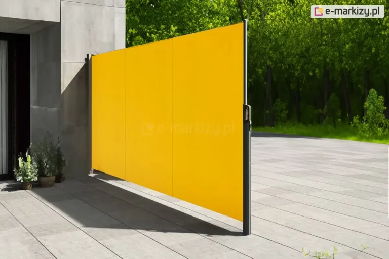 Zdjęcie prezentuje boczną markizę tarasową w formie rozwiniętego ekranu, który pełni funkcję osłony przeciwsłonecznej i parawanu zapewniającego prywatność na rozległym tarasie. Wyrazista tkanina w kolorze intensywnej, słonecznej żółci jest rozpięta pionowo, tworząc gładką płaszczyznę podzieloną subtelnymi pionowymi przeszyciami na trzy równe sekcje. Lewa krawędź osłony jest zamocowana do ciemnoszarej kasety przytwierdzonej do surowej, betonowej ściany nowoczesnego budynku. Prawa krawędź tkaniny kończy się aluminiowym profilem zintegrowanym z uchwytem, który jest wpięty w wolnostojący, ciemny słupek montażowy. Podłoże tarasu stanowią duże, jasnoszare płyty betonowe o gładkiej fakturze, na których widoczny jest wyraźny, wydłużony cień rzucany przez konstrukcję markizy. W tle, za linią tarasu, rozciąga się gęsty, soczyście zielony las lub park z wysokimi drzewami, co tworzy silny kontrast kolorystyczny z żółtym materiałem. Po lewej stronie, przy ścianie budynku, znajdują się dwie donice z niskimi, zielonymi roślinami ozdobnymi. Całość kompozycji jest skąpana w silnym, naturalnym świetle dziennym, podkreślającym nowoczesny i minimalistyczny charakter aranżacji przestrzeni zewnętrznej.