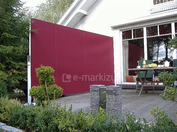 Parawan ogrodowy Bora Selt ustawiony przy ścianie białego budynku odgradza zejście z tarasu do ogrodu po lewej stronie budynku. Biała konstrukcja sprawia, że kaseta pozostaje niewidoczna, a słupek do mocowania tkaniny nie wyróżnia się z otoczenia szarego tarasu. Na tarasie osłoniętym czerwoną tkaniną parawanu ustawiono stolik ze śniadaniem i dwa krzesła z zielonymi poduszkami. W tle za stolikiem duże okno tarasowe. W otoczeniu tarasu wiele zieleni zapewnianej przez krzewy i drzewa.