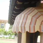 Zdjęcie przedstawia markizę koszową o półkolistym kształcie, zamontowaną nad oknem w jasnej elewacji budynku. Poszycie markizy wykonane jest z tkaniny w pionowe pasy w odcieniach beżu, szarości oraz intensywnego pomarańczu. Konstrukcja opiera się na białym stelażu, który nadaje jej wypukłą formę wachlarza. Dolna krawędź osłony wykończona jest dekoracyjną, falowaną falbaną z pomarańczową lamówką. Markiza jest przytwierdzona do ściany w kolorze piaskowym, tuż pod okapem dachu, gdzie widoczne są brązowe podbitki dachowe oraz ciemna rynna. W tle, za markizą, rozpościera się widok na nasłoneczniony ogród z zielonym trawnikiem i krzewami. W oddali widać fragment innego, jasnego budynku z dużymi przeszkleniami, w których odbija się światło słoneczne. Całość zdjęcia utrzymana jest w jasnej, ciepłej kolorystyce, podkreślającej ochronną funkcję markizy przed słońcem w upalny dzień.