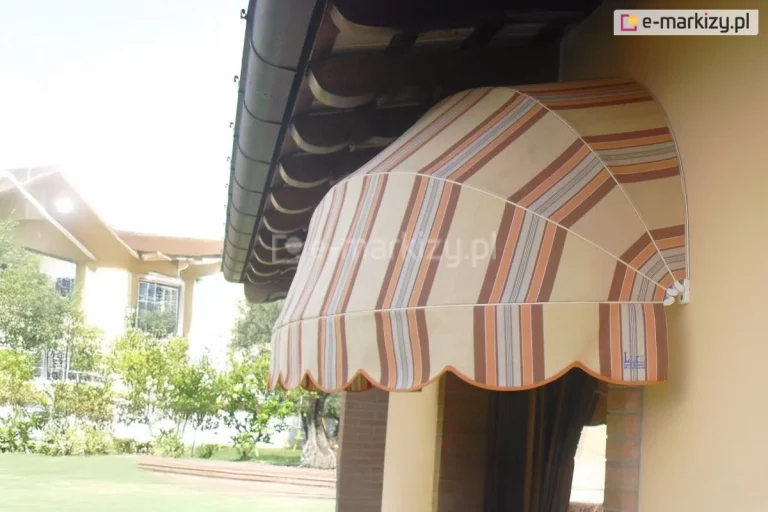 Zdjęcie przedstawia markizę koszową o półkolistym kształcie, zamontowaną nad oknem w jasnej elewacji budynku. Poszycie markizy wykonane jest z tkaniny w pionowe pasy w odcieniach beżu, szarości oraz intensywnego pomarańczu. Konstrukcja opiera się na białym stelażu, który nadaje jej wypukłą formę wachlarza. Dolna krawędź osłony wykończona jest dekoracyjną, falowaną falbaną z pomarańczową lamówką. Markiza jest przytwierdzona do ściany w kolorze piaskowym, tuż pod okapem dachu, gdzie widoczne są brązowe podbitki dachowe oraz ciemna rynna. W tle, za markizą, rozpościera się widok na nasłoneczniony ogród z zielonym trawnikiem i krzewami. W oddali widać fragment innego, jasnego budynku z dużymi przeszkleniami, w których odbija się światło słoneczne. Całość zdjęcia utrzymana jest w jasnej, ciepłej kolorystyce, podkreślającej ochronną funkcję markizy przed słońcem w upalny dzień.