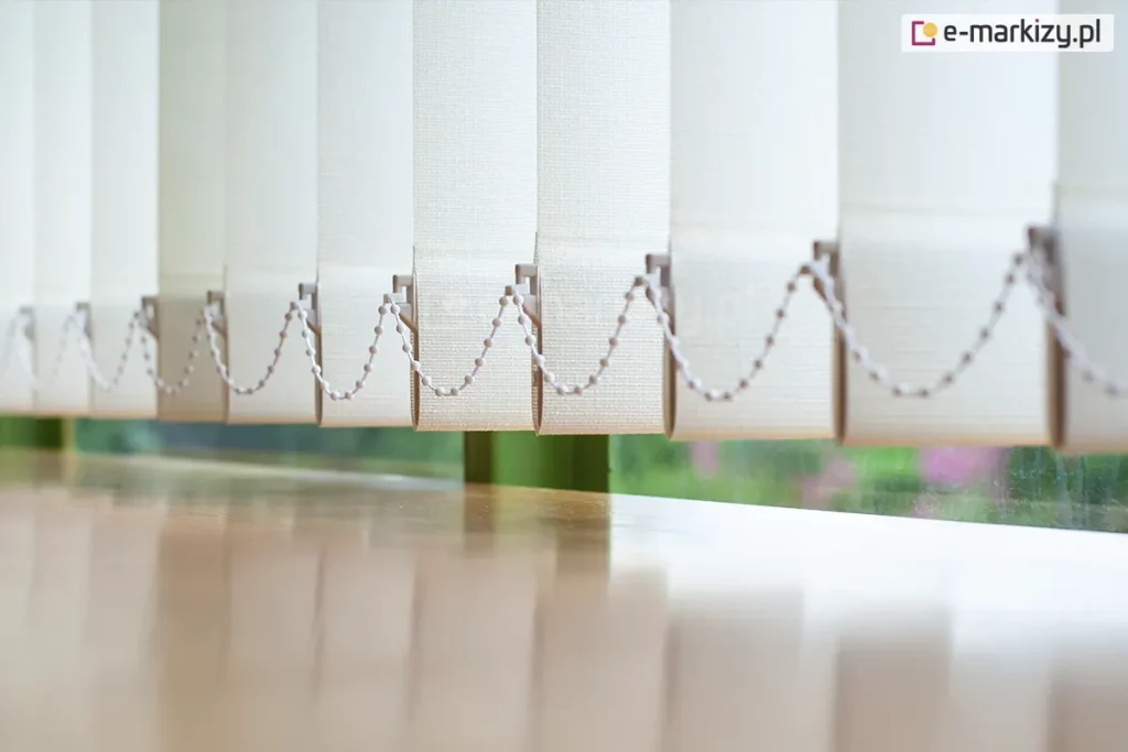 Zbliżenie na dolną część żaluzji pionowych, prezentujące mechanizm stabilizujący lamele nad powierzchnią jasnego, drewnianego blatu. Poszczególne pasy materiału w kolorze kremowym lub bardzo jasnego beżu mają wyraźną strukturę gęstego splotu tkaniny. W dolnej krawędzi każdej lameli znajduje się biały obciążnik, do którego wpięty jest plastikowy łańcuszek koralikowy. Łańcuszek ten łączy wszystkie elementy żaluzji pionowej, zapewniając ich równomierne ułożenie i zapobiegając nadmiernemu kołysaniu. Żaluzje pionowe są ustawione pod kątem, co tworzy rytmiczny, ząbkowany wzór na tle okna. Przez szybę widoczne jest rozmyte, zielone tło roślinności ogrodowej, co sugeruje montaż przesłony w pomieszczeniu z widokiem na naturę. Powierzchnia blatu pod żaluzjami jest gładka i lekko błyszcząca, odbijając pionowe pasy materiału oraz wpadające do wnętrza miękkie światło dzienne.