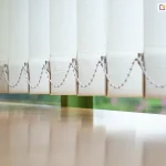 Zbliżenie na dolną część żaluzji pionowych, prezentujące mechanizm stabilizujący lamele nad powierzchnią jasnego, drewnianego blatu. Poszczególne pasy materiału w kolorze kremowym lub bardzo jasnego beżu mają wyraźną strukturę gęstego splotu tkaniny. W dolnej krawędzi każdej lameli znajduje się biały obciążnik, do którego wpięty jest plastikowy łańcuszek koralikowy. Łańcuszek ten łączy wszystkie elementy żaluzji pionowej, zapewniając ich równomierne ułożenie i zapobiegając nadmiernemu kołysaniu. Żaluzje pionowe są ustawione pod kątem, co tworzy rytmiczny, ząbkowany wzór na tle okna. Przez szybę widoczne jest rozmyte, zielone tło roślinności ogrodowej, co sugeruje montaż przesłony w pomieszczeniu z widokiem na naturę. Powierzchnia blatu pod żaluzjami jest gładka i lekko błyszcząca, odbijając pionowe pasy materiału oraz wpadające do wnętrza miękkie światło dzienne.